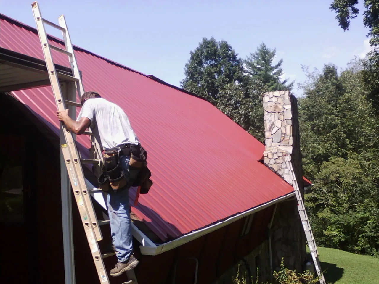 Roofer on a ladder beside a red metal roof and stone chimney — Soffit & Fascia Repair by Roofing Brothers in Wartrace